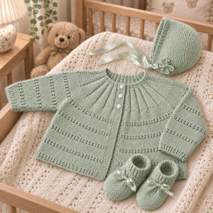 Könnte beinhalten: Ein salbeigrünes, gestricktes Baby-Set bestehend aus einer langärmeligen Strickjacke mit drei weißen Knöpfen, einer passenden Haube mit Satinband und kleiner Blumenapplikation sowie einem Paar Babyschuhen mit Satinbandschleifen.
