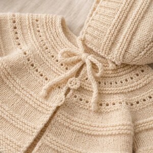 Könnte beinhalten: Eine beigefarbene Strickjacke mit passender Kapuze, die ein strukturiertes Rippenmuster, feine Lochmusterdetails entlang der Passe und einen zentralen Bindegürtel mit einem kleinen runden Knopf am Ausschnitt aufweist.