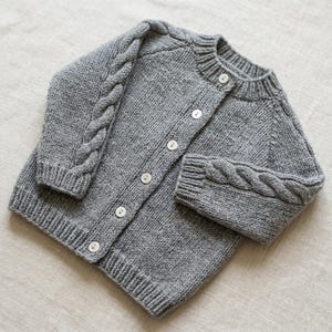 Può includere: Un cardigan in maglia grigia a maniche lunghe con un motivo a trecce sulle maniche, scollo rotondo a coste, polsini a coste e orlo a coste. La parte anteriore del capo è chiusa da sei piccoli bottoni rotondi bianchi lungo il centro.