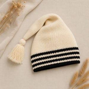 Könnte beinhalten: Eine cremefarbene Strickmütze mit einer langen, spitz zulaufenden Zipfelform, die in einer passenden cremefarbenen Quaste endet. Der untere Rand der Mütze ist mit drei markanten horizontalen schwarzen Streifen verziert, die einen Kontrast bilden.