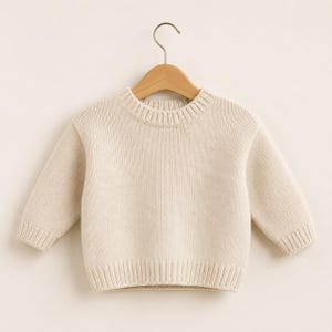 May include: A long-sleeved, cream-colored knit sweater featuring a ribbed crew neckline, ribbed cuffs, and a ribbed hem. The garment is displayed on a wooden hanger, showcasing a simple, solid-colored design with a consistent, fine-knit texture.