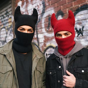 May include: Two ribbed knit balaclavas, one in solid black and one in solid red, each featuring two prominent, upright horn-like protrusions on the crown. The headwear covers the head and neck, leaving an opening for the eyes and nose area.