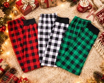 May include: Three pairs of lounge pants featuring a buffalo plaid pattern in red and black, white and black, and green and black. Each pair has a solid black elastic waistband and cuffed ankles, arranged side by side on a textured surface.