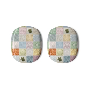 Op de afbeelding: Twee ovale toiletbrillen met een raster van vierkante tegels in pasteltinten zoals roze, blauw, groen, geel en oranje. Elke tegel toont reliëfpatronen, waaronder bloemmotieven, geometrische vormen, schelpen en groene zeeschildpadden.