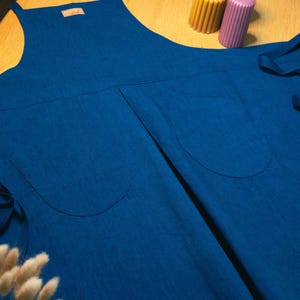 Puede incluir: Un delantal azul profundo con un peto central, dos grandes bolsillos delanteros y largas cintas de tela en los laterales. Una pequeña etiqueta rectangular está cosida en la parte superior del pecho del delantal extendido sobre madera.