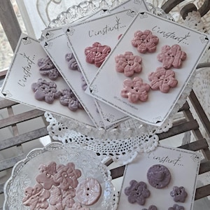 Può includere: Una collezione di bottoni in ceramica in varie tonalità di malva, lavanda e rosa polvere, a forma di fiori e cerchi. I bottoni sono esposti su cartoncini rettangolari bianchi con il testo l'instant... e all'interno di una ciotola di vetro.