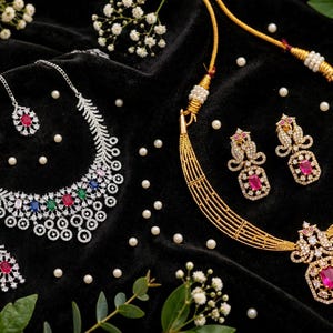 May include: A silver-toned necklace and earring set with multi-colored stones and a gold-toned necklace and earring set featuring pink rectangular stones, both adorned with clear crystals and arranged on a black fabric surface with white pearls.