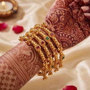May include: A set of four gold-toned bangles featuring a twisted rope texture, small dangling charms, and alternating square-cut red and green stones. The bangles are arranged in a stack, showcasing a metallic finish and intricate detailing.