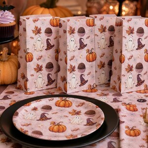 Puede incluir: Un conjunto de bolsas de papel rosa claro y un plato de papel a juego con un patrón repetitivo de Halloween de fantasmas blancos, calabazas naranjas, sombreros de bruja morados, trufas marrones y hojas de otoño sobre un camino de mesa.