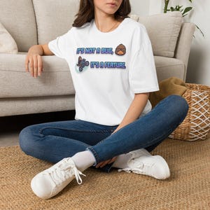 Peut inclure: Un t-shirt blanc à manches courtes avec une impression graphique portant le texte "IT'S NOT A BUG, IT'S A FEATURE." Le motif comprend une petite créature brune portant des lunettes et une seconde illustration de la même créature à moto.