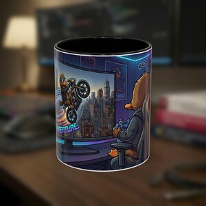 May include: A ceramic mug with a black interior and a graphic print of a duck sitting in a gaming chair, holding a controller and watching a screen that displays another duck performing a motorcycle stunt in a city with the text RAD and A FEATURE.