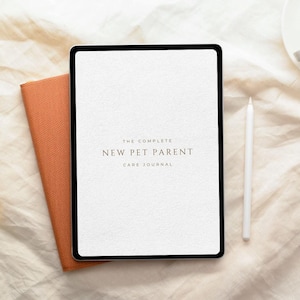 May include: A digital tablet displaying a white screen with the text THE COMPLETE NEW PET PARENT CARE JOURNAL in black font. Beside the tablet is a white stylus and a textured orange notebook cover, all arranged on a light fabric surface.