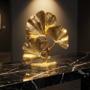 Puede incluir: Una escultura de metal dorado que presenta tres hojas de ginkgo biloba unidas a un tallo central. Las hojas tienen una superficie texturizada y estriada, montadas sobre una base rectangular con un acabado fino, granulado y brillante.