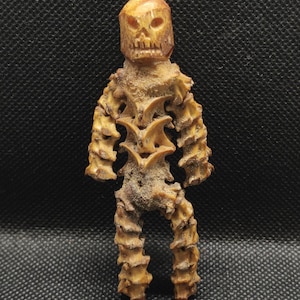 May include: A tan-colored, humanoid figurine crafted from textured, organic-looking material. The figure features a carved skull-like head with hollow eye sockets and a jagged mouth, and a body constructed from stacked, vertebrae-like segments.