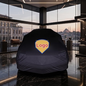 Pode incluir: Uma capa de carro de cor escura sobre um veículo, apresentando um emblema central em forma de escudo com fundo amarelo, borda azul e o texto vermelho Logo impresso no centro, posicionado num showroom moderno com paredes de vidro.