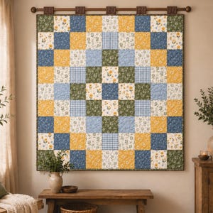 Puede incluir: Una colcha de patchwork cuadrada con una cuadrícula de cuadrados de tela alternos en amarillo mostaza, verde salvia, azul polvoriento y blanco con patrones florales o de cuadros vichy. La colcha cuelga de una barra de madera oscura.