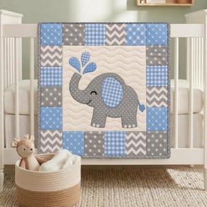 Könnte beinhalten: Eine quadratische Babydecke mit einem cremefarbenen Mittelteil, auf dem ein grauer Elefant appliziert ist, der blaues Wasser sprüht. Der Rand besteht aus einem Patchwork-Muster aus blauen und grauen Quadraten mit Stern-, Zickzack- und Karomustern.