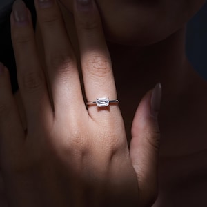 Puede incluir: Un anillo de tono plateado con una gema transparente rectangular de corte esmeralda, montada horizontalmente sobre una banda de metal fina y pulida. La piedra muestra facetas claras que reflejan la luz, creando un punto focal brillante.