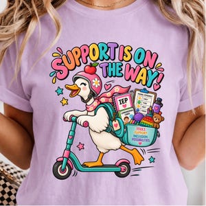 May include: A light purple t-shirt featuring a graphic of a white duck wearing a pink helmet and scarf, riding a teal scooter. The duck carries a backpack filled with school supplies, including an IEP folder, visual schedule, and teddy bear.