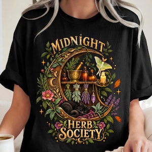 Puede incluir: Una camiseta negra con un gráfico de una luna creciente que contiene estantes con un mortero, botellas de vidrio, una vela y un cristal colgante. Un gato negro duerme debajo, rodeado de flores, hierbas y una polilla, con el texto MIDNIGHT HERB SOCIETY.