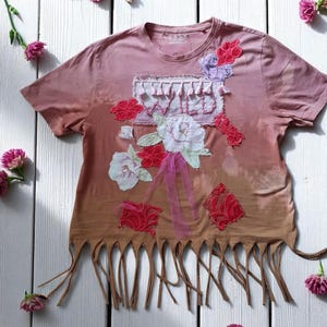May include: A pink and brown tie-dye t-shirt with the word "WILD" embroidered on the front. The shirt is decorated with floral appliqués in red, white, and purple, and has a fringe hem.