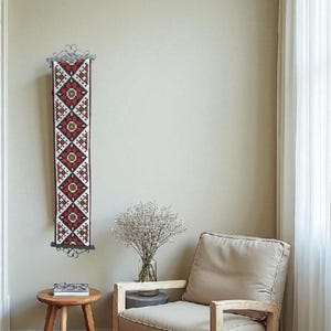 Könnte beinhalten: Ein langer, rechteckiger Wandbehang mit einem geometrischen Muster in Rot, Weiß und Schwarz. Der Stoff ist an einer dekorativen Metallstange befestigt. Der Wandbehang wird in einem Raum mit neutralen Wänden und Möbeln ausgestellt.