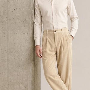 Puede incluir: Una camisa de manga larga color crema con cuello en punta y puños, combinada con pantalones de pana beige de talle alto con pliegues delanteros, cierre de botón y corte ancho, junto con mocasines de cuero marrón oscuro.