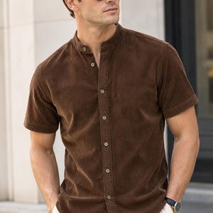 May include: A short-sleeved brown corduroy shirt featuring a band collar, a full-length front button placket with matching brown buttons, and a textured vertical ribbed fabric finish. The shirt has a straight hemline and cuffed sleeves.