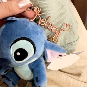 Puede incluir: Un llavero de peluche azul con una etiqueta de alambre en tono oro rosa que dice Kelsey en letra cursiva, con un pequeño detalle de corazón al final. El peluche tiene ojos negros grandes, detalles en azul claro y orejas de color rosa.