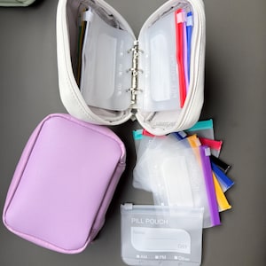 Pode incluir: Um fichário branco com zíper e mecanismo de argolas de metal contendo bolsas plásticas translúcidas, ao lado de um estojo lilás claro com zíper e várias bolsas translúcidas soltas rotuladas como PILL POUCH com campos para nome, dia, AM, PM e Other.