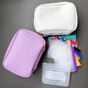 Op de afbeelding: Een wit rechthoekig tasje met rits en een lavendelkleurig rechthoekig tasje met rits. Daarnaast liggen verschillende doorzichtige plastic pillenzakjes met gekleurde ritssluitingen, elk bedrukt met de tekst PILL POUCH, Name, Day, AM, PM, Other.