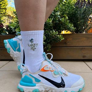 Op de afbeelding: Een witte enkelsok met een geborduurde blauwe bloem en de tekst Lucky IVF SOCKS met een klein hartje, gedragen met een witte sportschoen met een zwarte swoosh, felblauwe accenten en een gestructureerde witte rubberen zool.