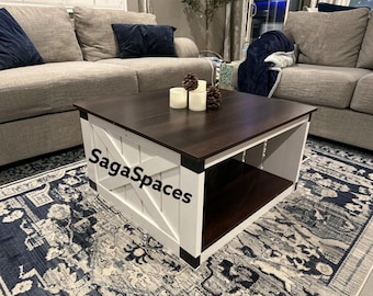 May include: A square coffee table with a dark brown wooden top and a white base featuring a cross-brace design on the sides. The table has an open lower shelf and the text SagaSpaces is printed in black on the front white panel.