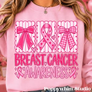 Op de afbeelding: Een lichtroze sweatshirt met een afbeelding van drie verticale panelen met een effen roze strik, een roze bewustzijnslint en een roze geruite strik. Daaronder staat de tekst BREAST CANCER AWARENESS in het roze, omringd door kleine hartjes.