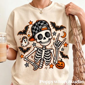 May include: A beige crewneck sweatshirt featuring a graphic of a skeleton wearing a black and gray checkered baseball cap. The design includes a small ghost, several bats, orange stars, gray stars, and a jack-o'-lantern pumpkin.