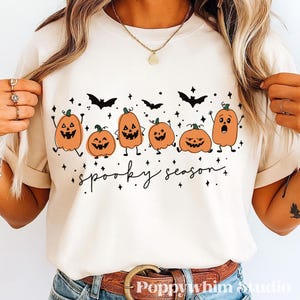 May include: A cream-colored t-shirt featuring a graphic print of six orange jack-o'-lanterns with various facial expressions, black bats, and small stars. Below the pumpkins, the text "spooky season" is written in a black cursive script font.