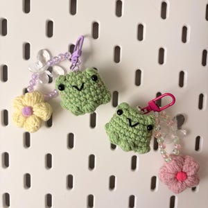 May include: Two crochet frog keychains featuring green yarn bodies with black bead eyes and stitched mouths. Each keychain includes a colorful plastic clasp, a string of translucent beads, a clear butterfly charm, and a crocheted flower.