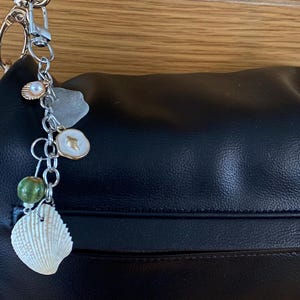 Puede incluir: Un colgante de metal plateado para bolso que incluye una concha blanca, una cuenta verde, un pequeño dije de esmalte blanco, un trozo de vidrio marino esmerilado y una concha pequeña con una perla, unido a un bolso de cuero negro.