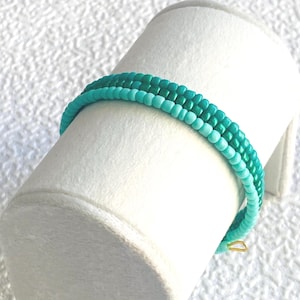 Könnte beinhalten: Ein mehrreihiges Armband aus kleinen, runden Glasperlen in verschiedenen Türkis-, Blaugrün- und Hellblautönen. Das Armband hat eine gleichmäßige Perlengröße und wird mit einem kleinen, dünnen Verschluss aus goldfarbenem Metall geschlossen.
