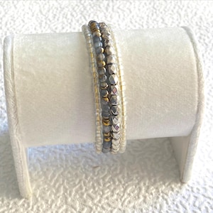 Könnte beinhalten: Ein mehrreihiges Armband bestehend aus kleinen klaren Glasperlen, metallisch goldfarbenen Perlen, glatten grauen Perlen und facettierten silberfarbenen Metallperlen, die in einem geschichteten Design auf einem weißen Ständer angeordnet sind.