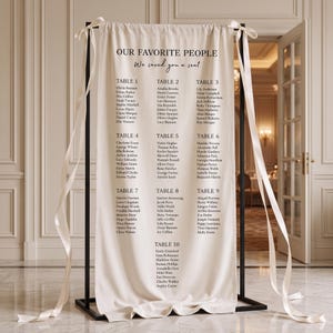 May include: A cream-coloured fabric wedding seating chart hanging on a black metal frame. The text reads OUR FAVORITE PEOPLE We saved you a seat, followed by lists of names organised into ten tables. Long white ribbons are tied to the top corners.