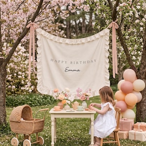 May include: A cream-colored fabric banner with ruffled edges, featuring the black text "HAPPY BIRTHDAY" and "Emma" in script. The banner is suspended by pink ribbons between two trees, creating a backdrop for a small table setting.