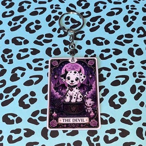 Peut inclure: Un porte-clés rectangulaire présentant une illustration de carte de tarot intitulée THE DEVIL. Le motif représente un chiot tacheté avec de petites cornes et des ailes de chauve-souris assis sur un piédestal, entouré de roses violettes.