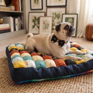 May include: A fawn-colored pug wearing a black bow tie with rainbow stripes, resting on a rectangular pet bed. The bed features a quilted top made of small, multi-colored fabric squares in shades of yellow, orange, teal, and black, with a solid navy border.