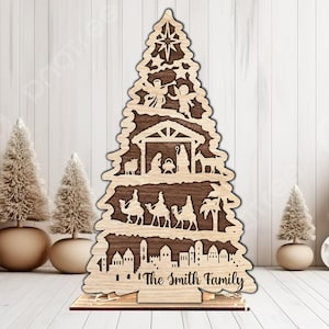 Könnte beinhalten: Ein lasergeschnittener Weihnachtsbaum aus Holz mit einer mehrschichtigen Krippenszene, einem Stern an der Spitze, Engeln, der heiligen Familie, Kamelen und einer Stadtsilhouette. Der Sockel ist mit dem Text The Smith Family in schwarzer Schrift graviert.