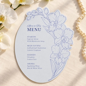 May include: An oval-shaped menu card with blue text and a blue floral line drawing on the right side. The text reads: Alexa & Tony MENU, STARTER Caprese Salad Wild Mushroom Soup, MAIN COURSE Grilled Beef Tenderloin Lobster Linguine, DESSERT Tiramisu Lemon Tart, DRINKS Sparkling Wine Red & White Wine.