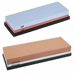 May include: Two rectangular sharpening stones set in black bases. The top stone is light blue and reddish-brown with 800 and 240 grit markings. The bottom stone is tan and white with 3000# and 8000# grit markings printed on the side surfaces.