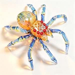 Op de afbeelding: Een glazen spinnenfiguur met een doorschijnend oranje en geel bolvormig achterlijf met een honingraatstructuur, een bijpassend borststuk, acht gesegmenteerde blauwe en gele poten en acht kleine, felrode ronde ogen op de kop.