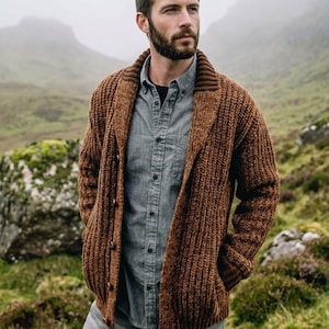 May include: A chunky knit brown cardigan with a wide shawl collar and vertical ribbed texture, worn over a grey button-down shirt and grey trousers. The cardigan features a front button closure with dark brown buttons and long sleeves.