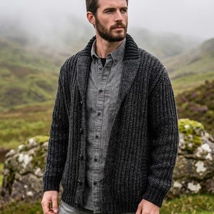 Könnte beinhalten: Eine grob gestrickte Strickjacke in Anthrazit mit Schalkragen und Knopfleiste auf der Vorderseite. Das Kleidungsstück wird über einem grauen Hemd und einer hellgrauen Hose getragen und weist eine dicke Rippenstruktur im gesamten Strick auf.